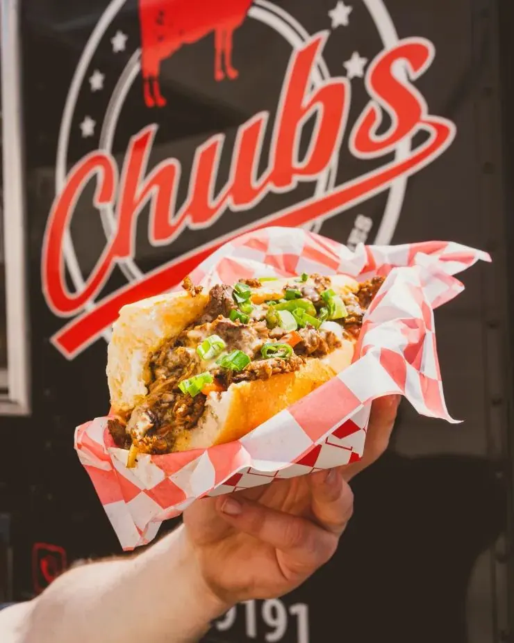 Philly cheesesteak in checkered paper, held in front of the Chubs truck